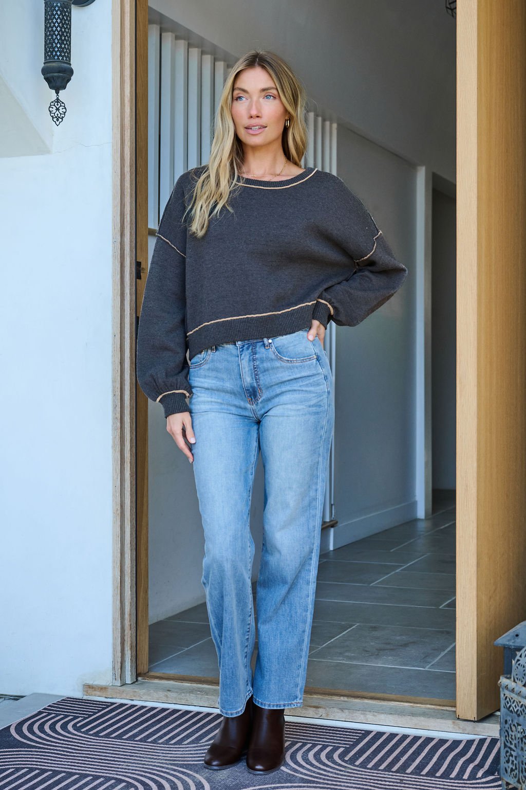 A woman stands in a doorway wearing a Charcoal Contrast Seam Detail Pullover, light blue jeans, and brown ankle boots.