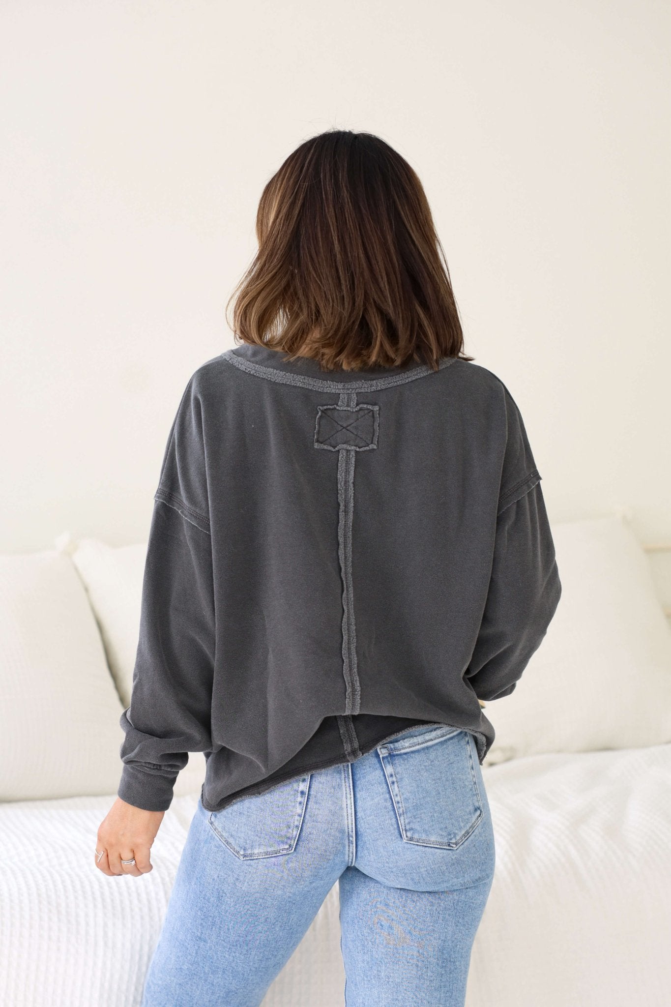 A woman with shoulder-length brown hair wears a cozy Charcoal Exposed Seam Detail Sweatshirt and light blue jeans in a bright room.