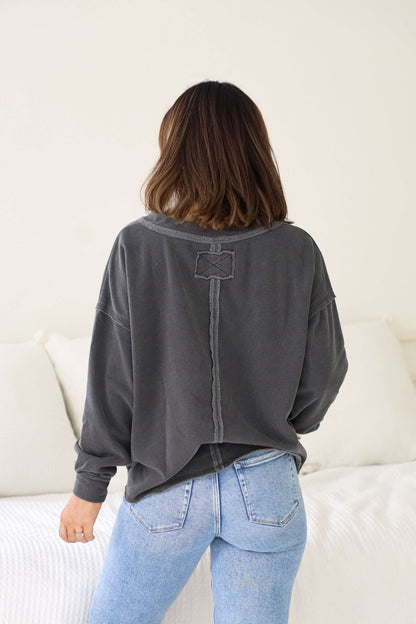 A woman with shoulder-length brown hair wears a cozy Charcoal Exposed Seam Detail Sweatshirt and light blue jeans in a bright room.