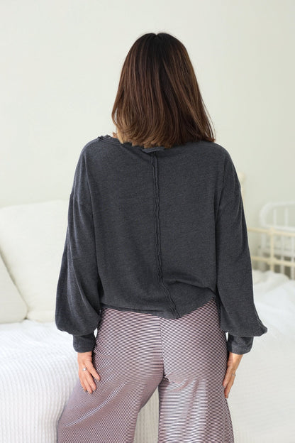 A woman with brown hair wears a Charcoal Mineral Wash Long Sleeve Top - FINAL SALE and striped pants in a bright bedroom.
