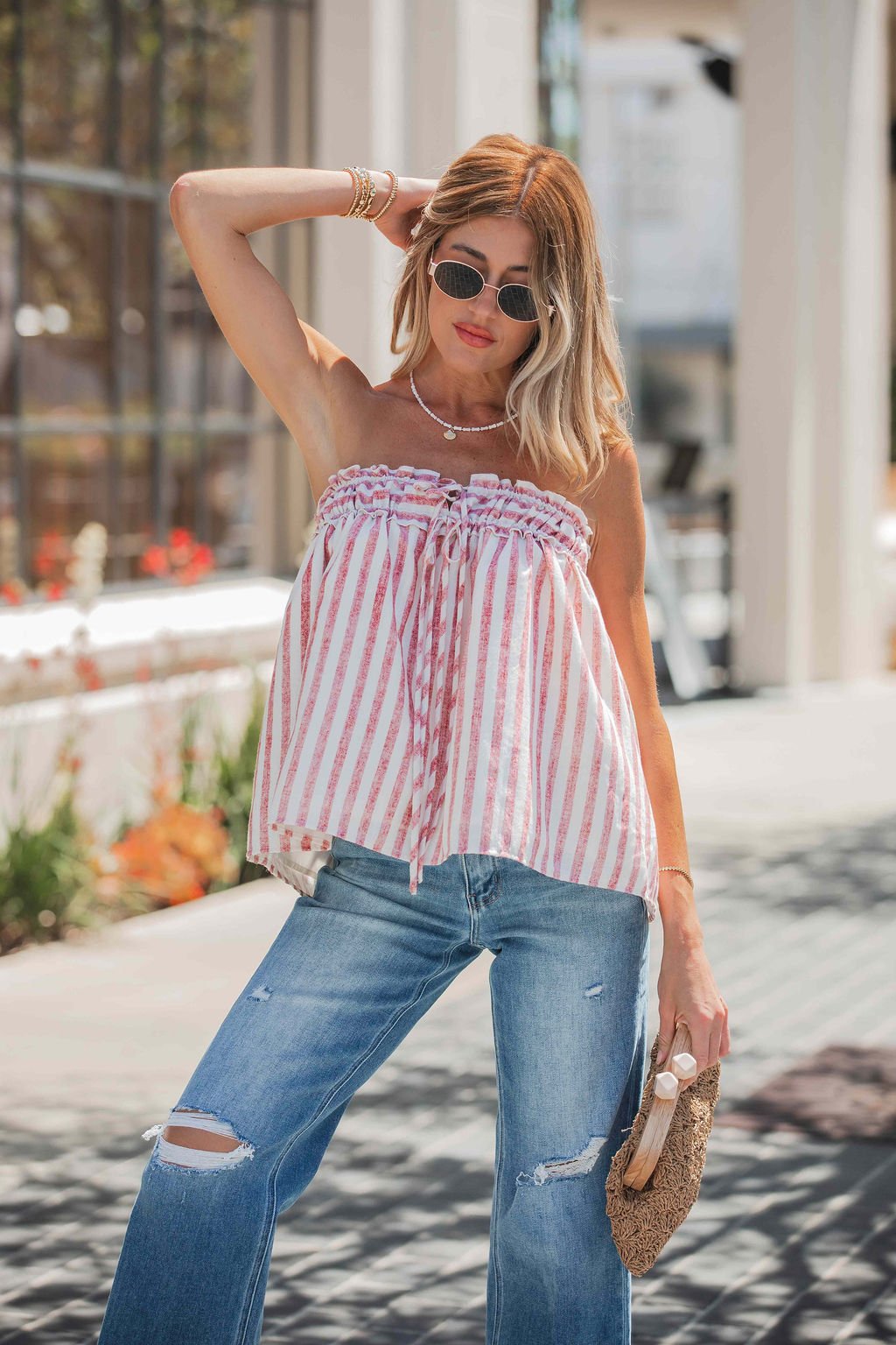 Woman in sunglasses and ripped jeans poses outdoors in the Cherry Lane Strapless Stripe Top, holding a straw clutch.