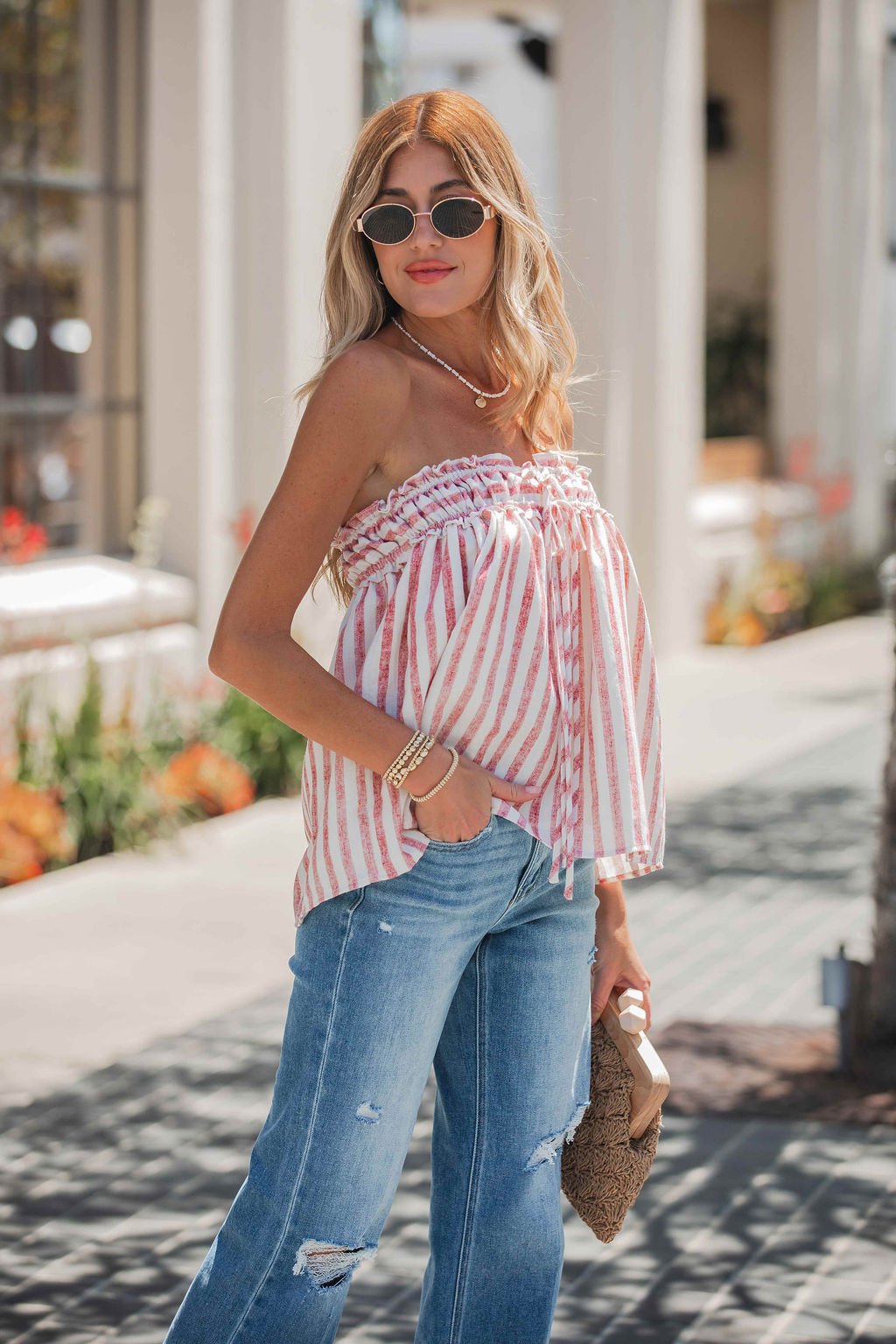 Woman in sunglasses wearing the Cherry Lane Strapless Stripe Top and blue jeans, holding a brown woven clutch outdoors.