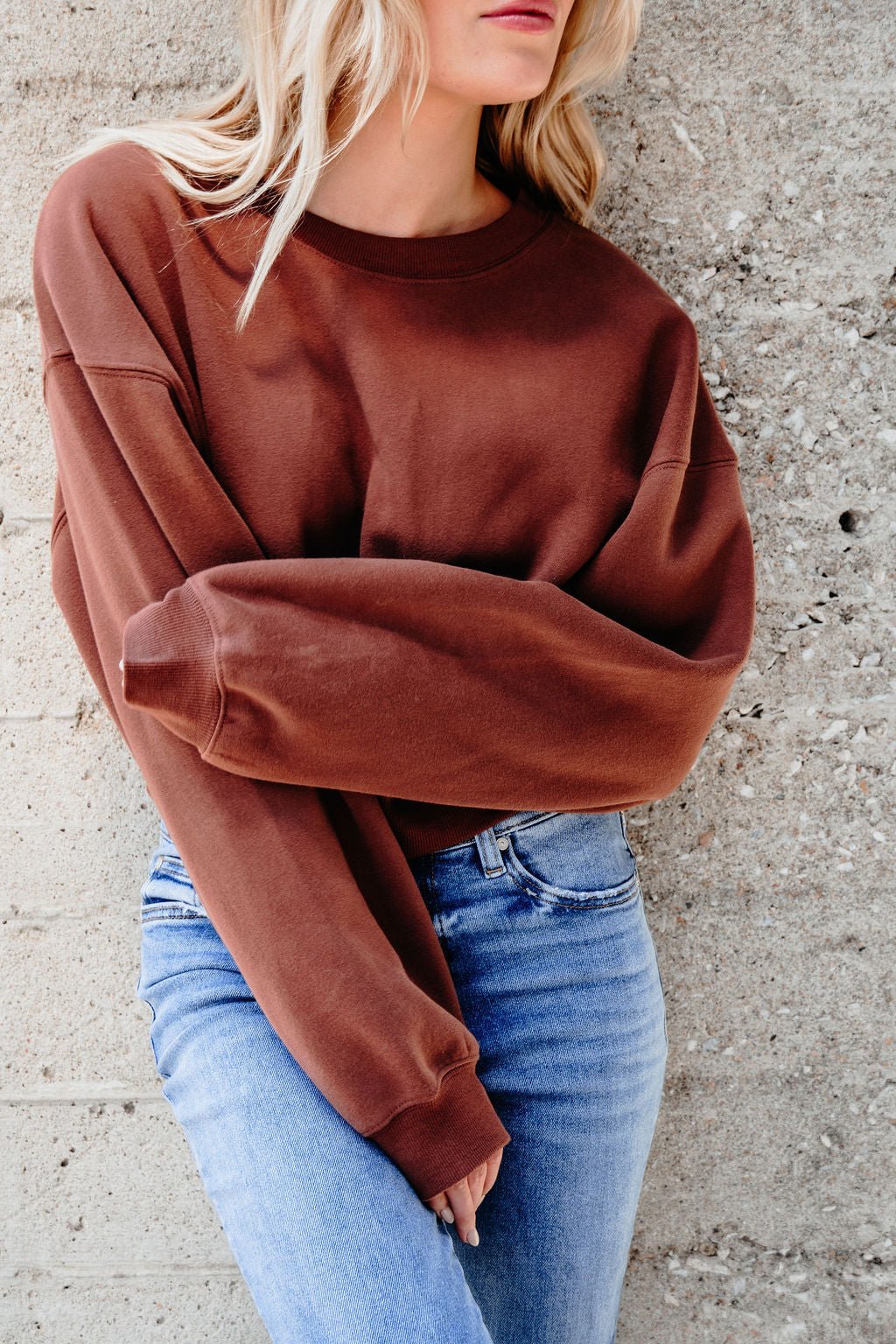 A woman in a Chestnut Crew Neck Ribbed Sweatshirt and light blue jeans stands against a textured concrete wall, face partially out of frame.