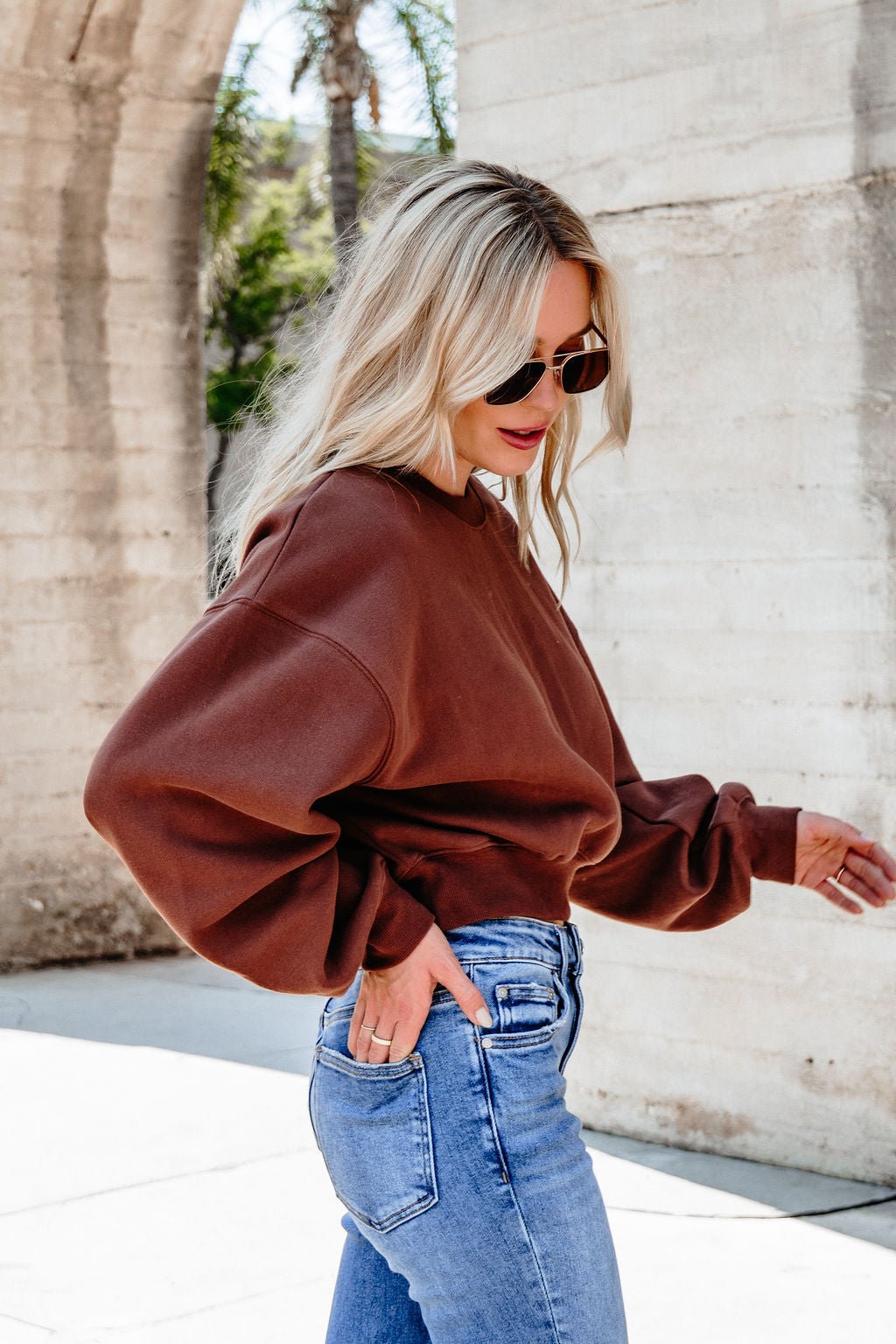 Woman with long blonde hair wears a Chestnut Crew Neck Ribbed Sweatshirt, blue jeans, and sunglasses by a concrete wall outdoors.