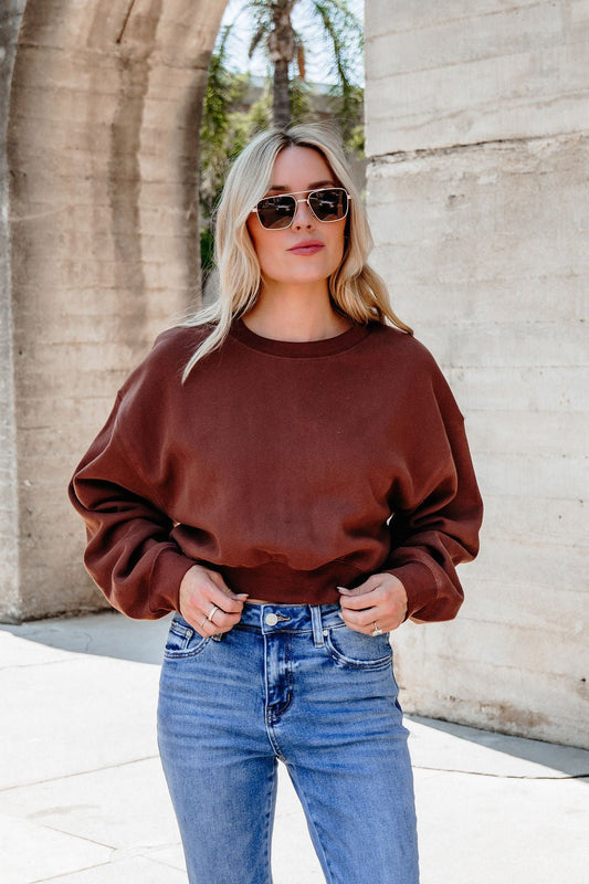 A woman in sunglasses, blue jeans, and a Chestnut Crew Neck Ribbed Sweatshirt stands outdoors by a stone archway.