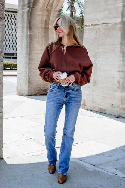 A woman in a Chestnut Crew Neck Ribbed Sweatshirt, blue jeans, and sunglasses smiles outside holding a coffee cup.