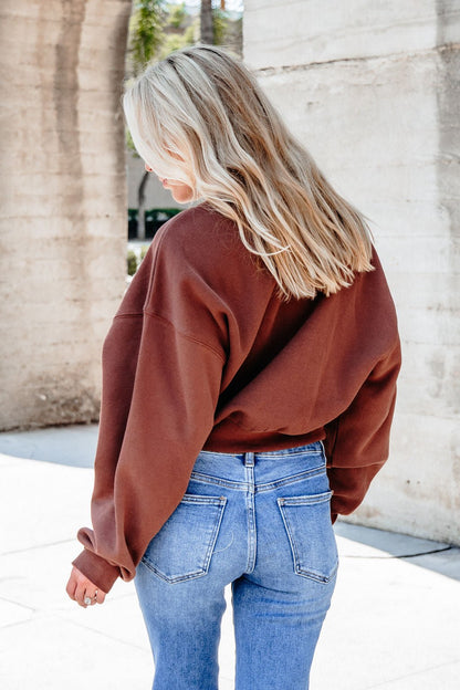 A woman with blonde hair wears a Chestnut Crew Neck Ribbed Sweatshirt and blue jeans, standing outdoors facing away.