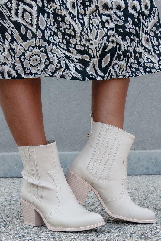 A person stands on a textured surface in a patterned skirt and Chinese Laundry Beige U See Ankle Western Booties – FINAL SALE.