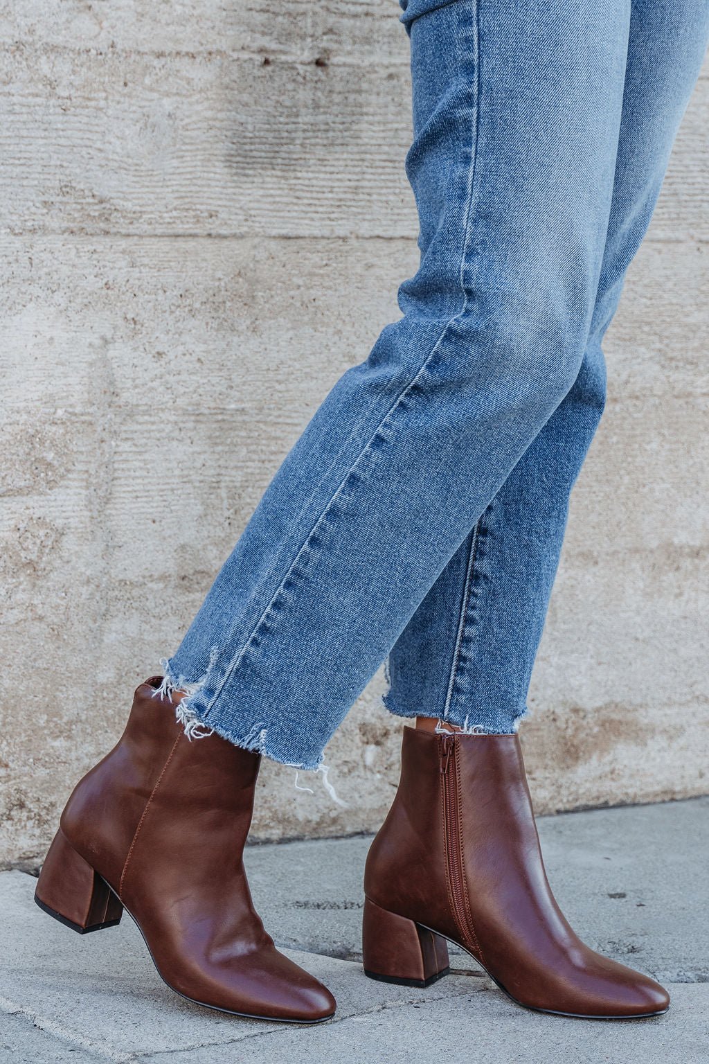 Wearing cropped blue jeans and Chinese Laundry Davinna Ankle Booties, a person stands on a sidewalk against a beige wall.