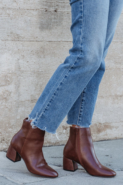 Wearing cropped blue jeans and Chinese Laundry Davinna Ankle Booties, a person stands on a sidewalk against a beige wall.