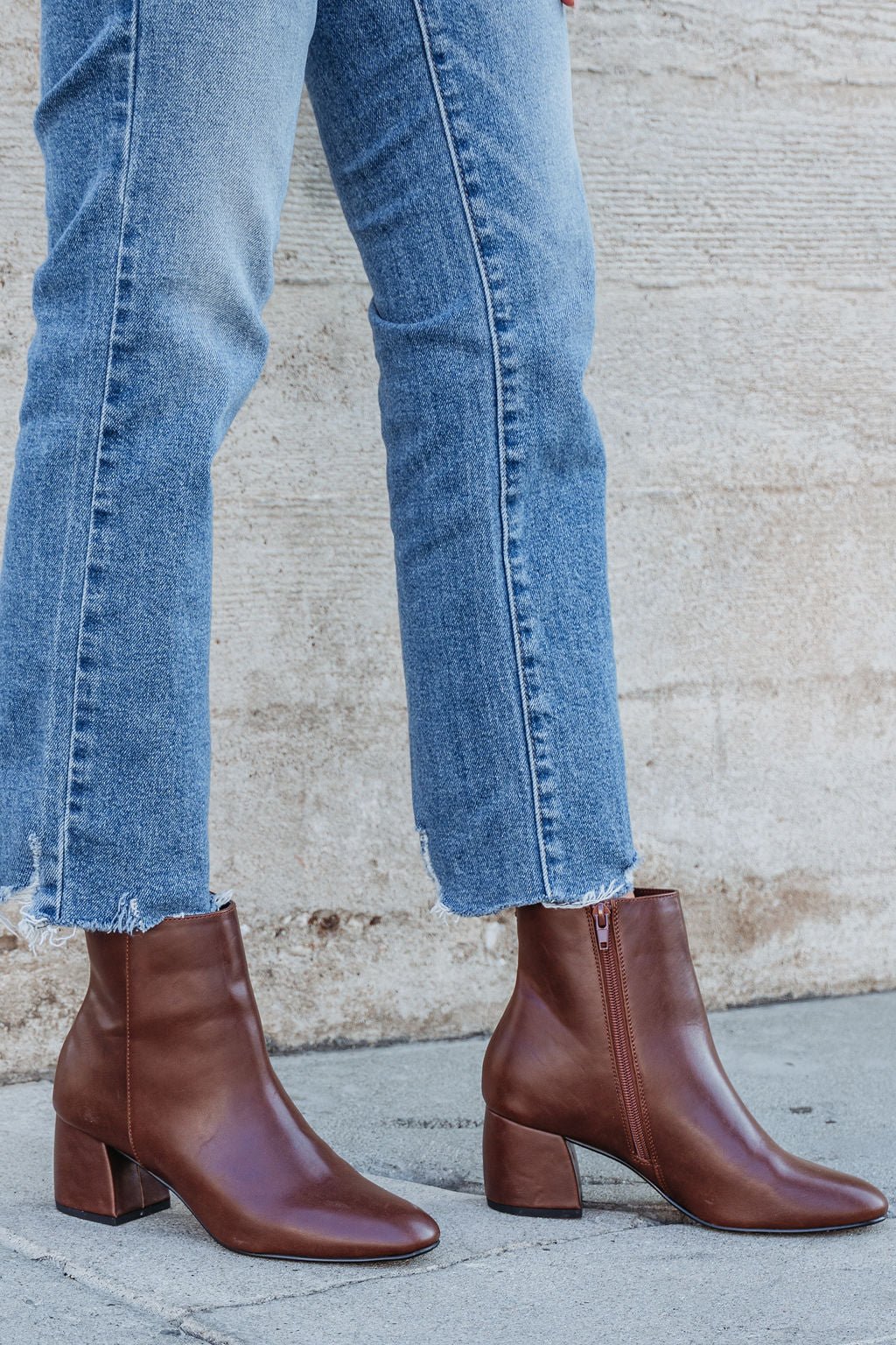 Person in blue frayed-hem jeans and Chinese Laundry Davinna Ankle Booties stands on a sidewalk by a beige wall.