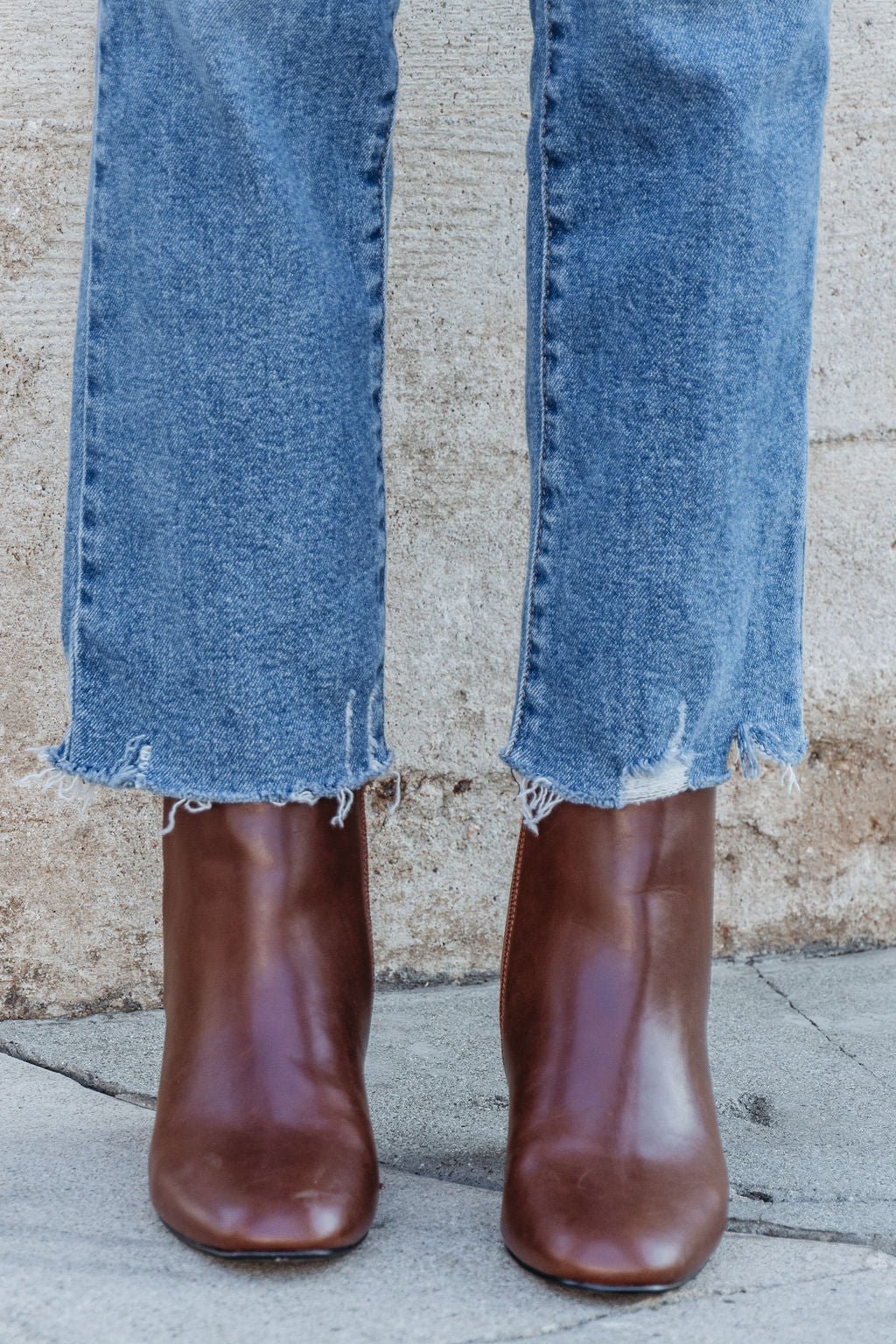 Person in frayed hem blue jeans and Chinese Laundry Davinna Ankle Booties stands on a sidewalk by a stone wall.