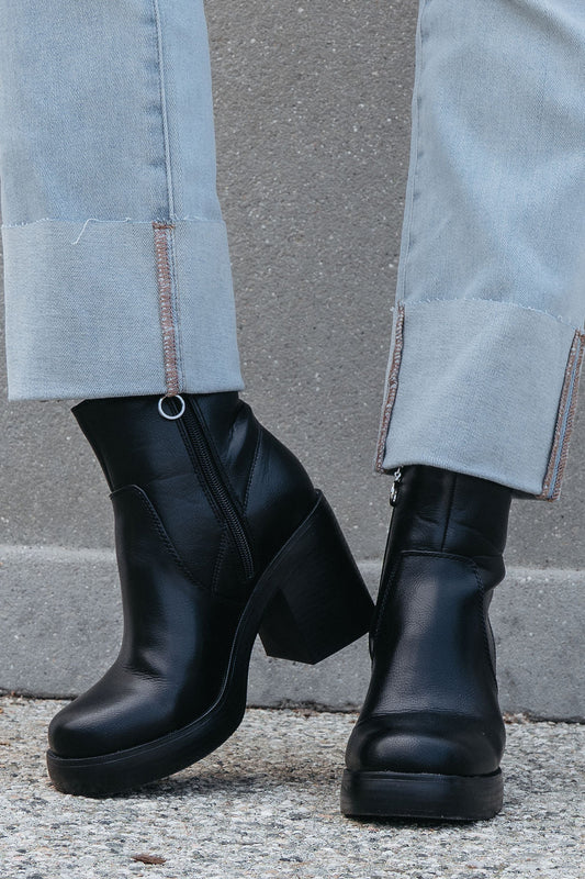 Someone wearing Chinese Laundry Groovy Black Heeled Ankle Boots and frayed light-wash jeans stands on concrete.