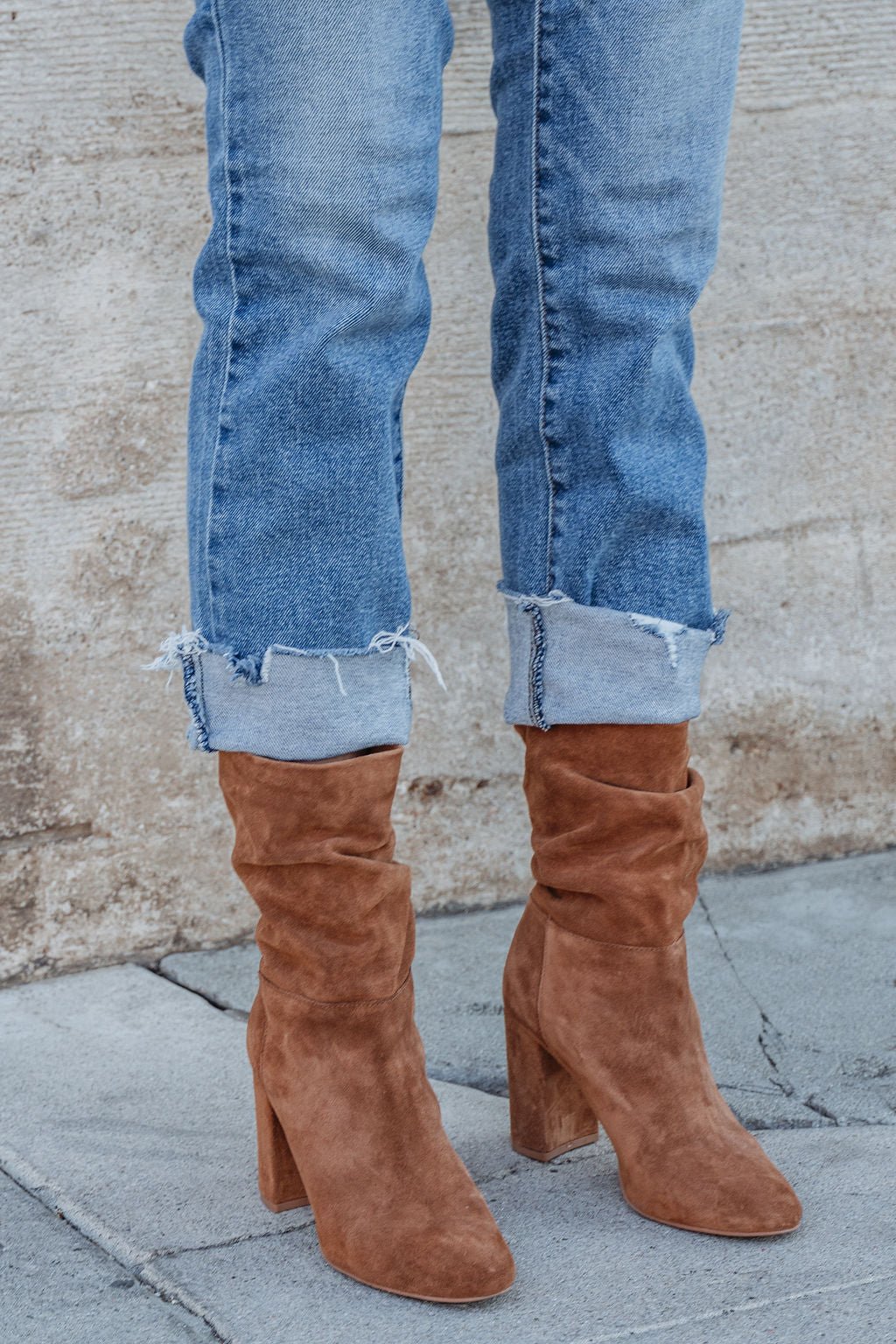 Wearing light blue cuffed jeans and Chinese Laundry Kipper Brown Suede Heeled Boots, standing by a textured wall on gray pavement.