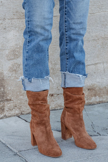 Wearing light blue cuffed jeans and Chinese Laundry Kipper Brown Suede Heeled Boots, standing by a textured wall on gray pavement.