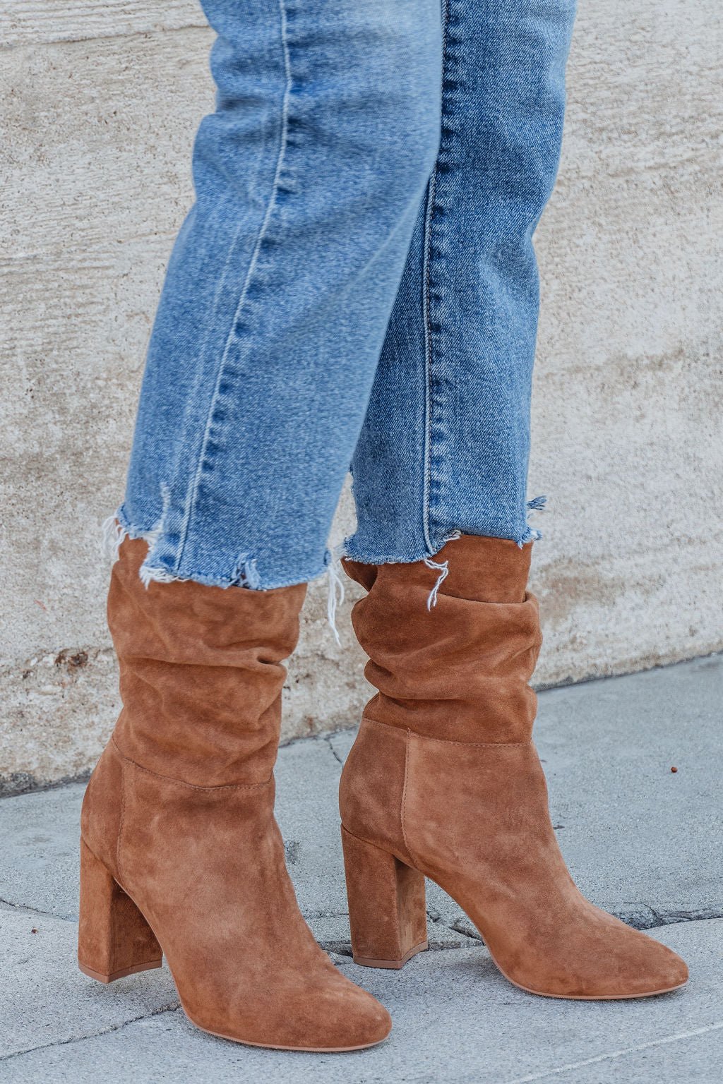 Person in frayed blue jeans and Chinese Laundry Kipper Brown Suede Heeled Boots, standing on a sidewalk—a fall must-have.