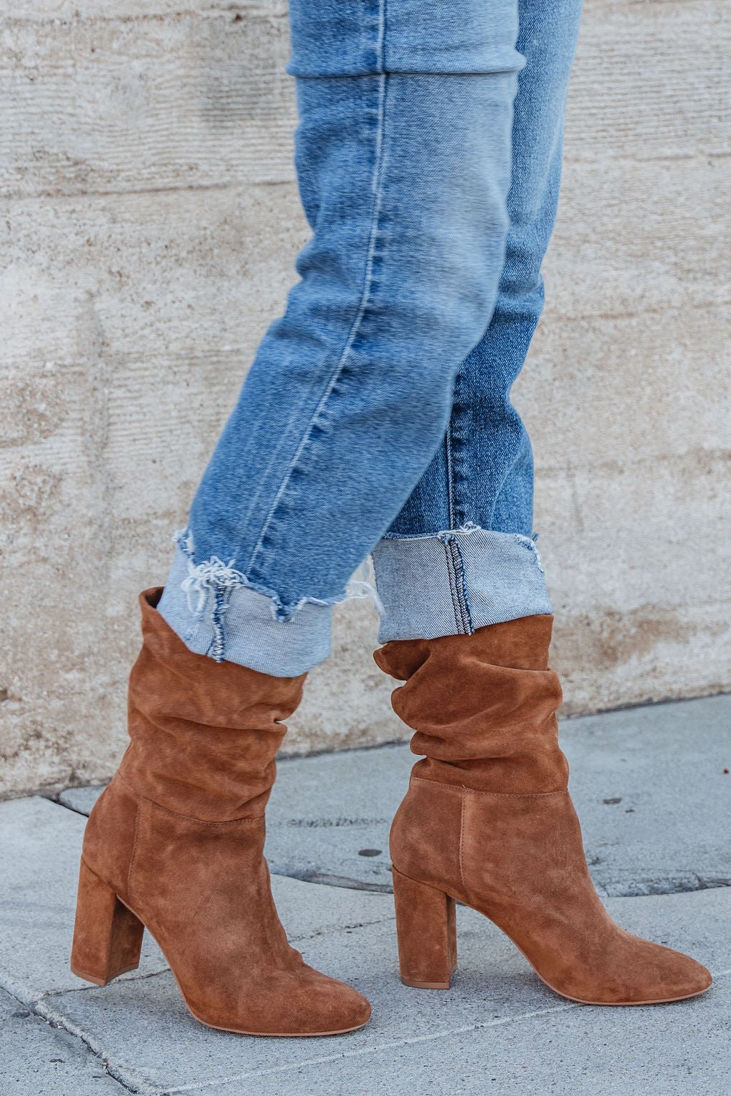 A person in cuffed blue jeans and Chinese Laundry Kipper Brown Suede Heeled Boots stands on a concrete sidewalk—a fall essential.
