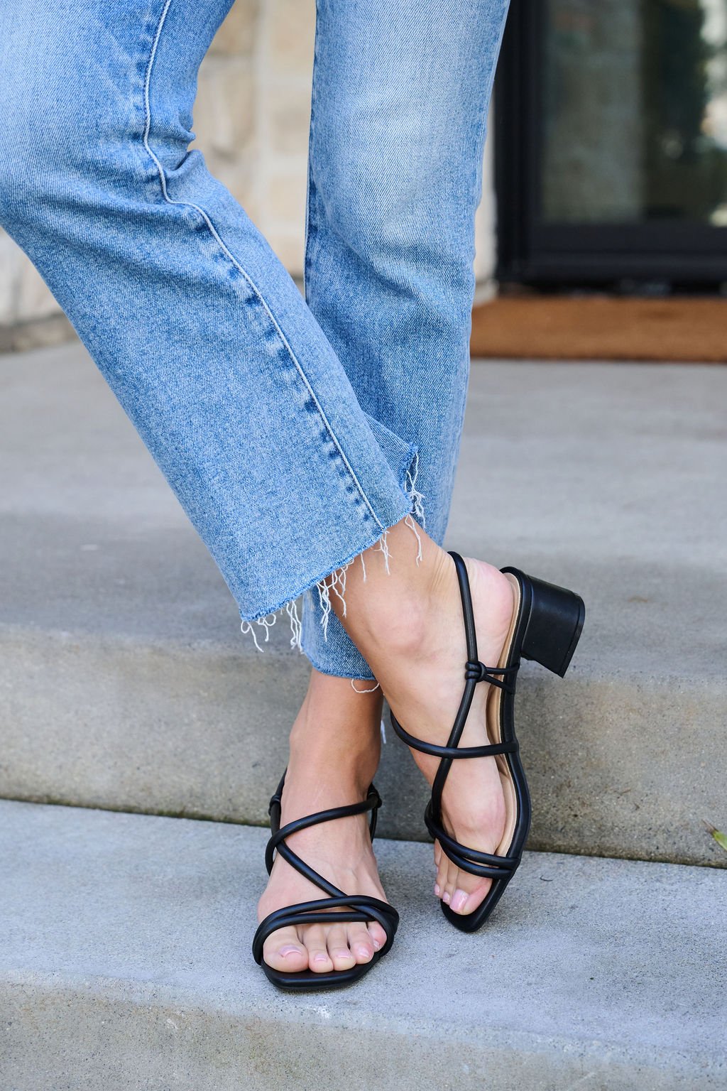 Wearing frayed blue jeans and Chinese Laundry Lighten Black Strappy Heeled Sandals, standing on outdoor steps.