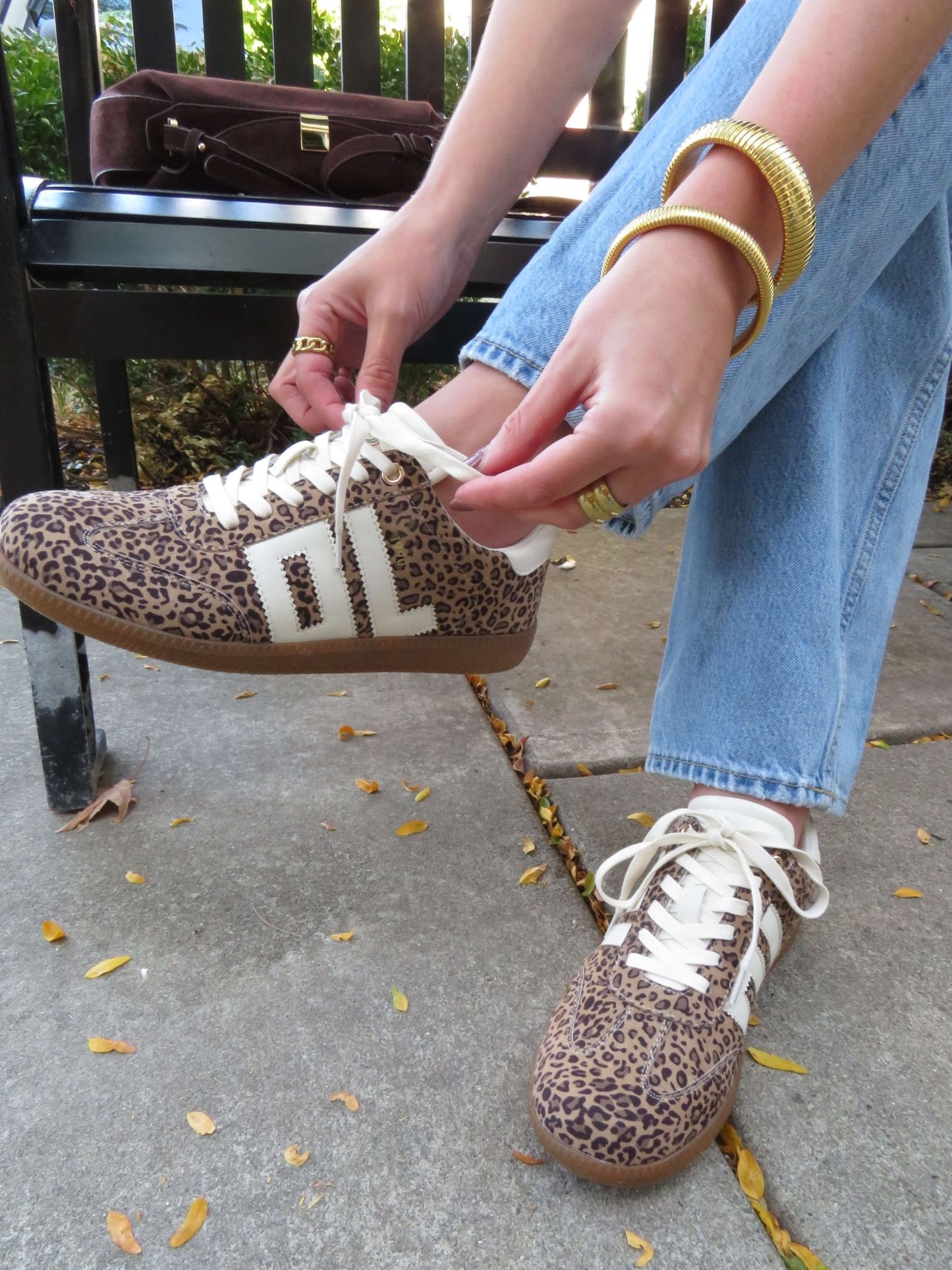 Wearing Chinese Laundry Trackstar Leopard Print Sneakers - FINAL SALE and jeans, a person ties their shoe on the sidewalk with gold jewelry.