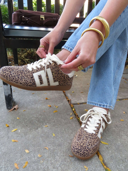 Wearing Chinese Laundry Trackstar Leopard Print Sneakers - FINAL SALE and jeans, a person ties their shoe on the sidewalk with gold jewelry.