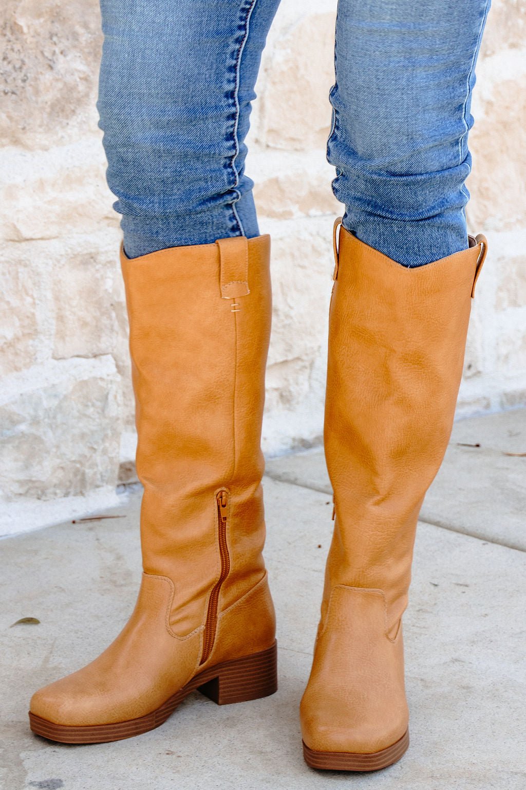 Person wearing fitted blue jeans tucked into Chinese Laundry Yurie Tan Knee High Boots, standing on a concrete surface by a stone wall.