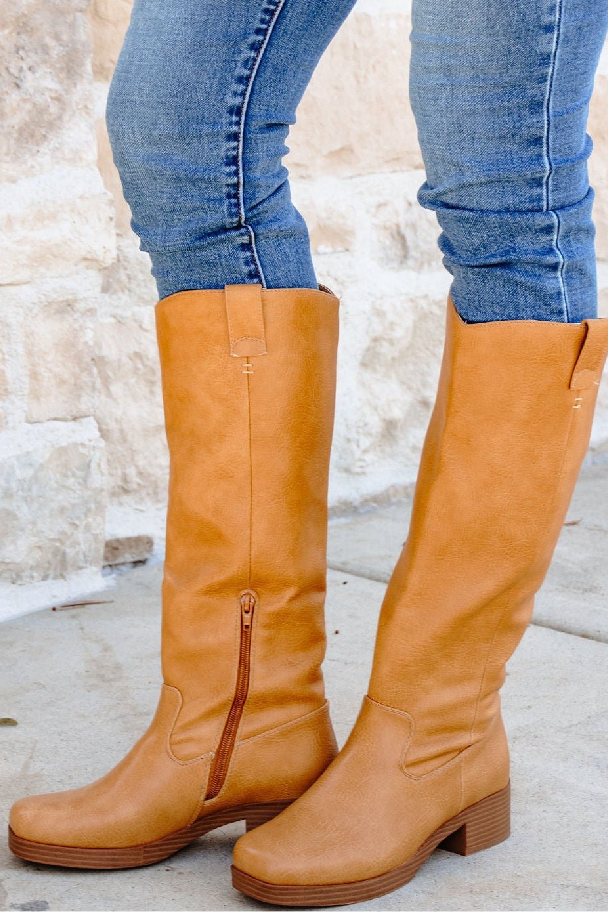 Tan knee-high boots worn with blue jeans against a stone wall background