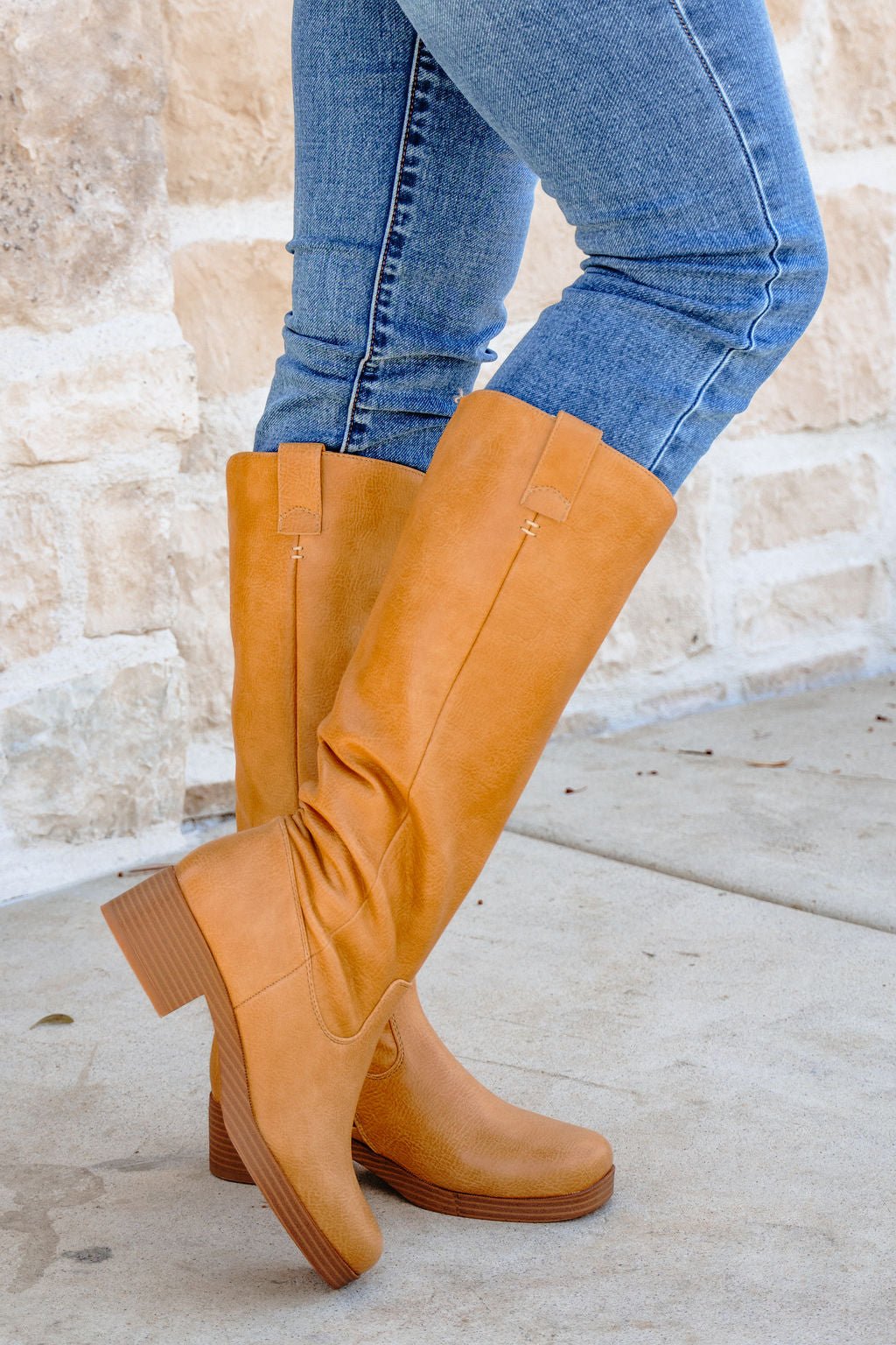 Person wearing blue jeans and Chinese Laundry Yurie Tan Knee High Boots, standing on a concrete sidewalk by a stone wall.