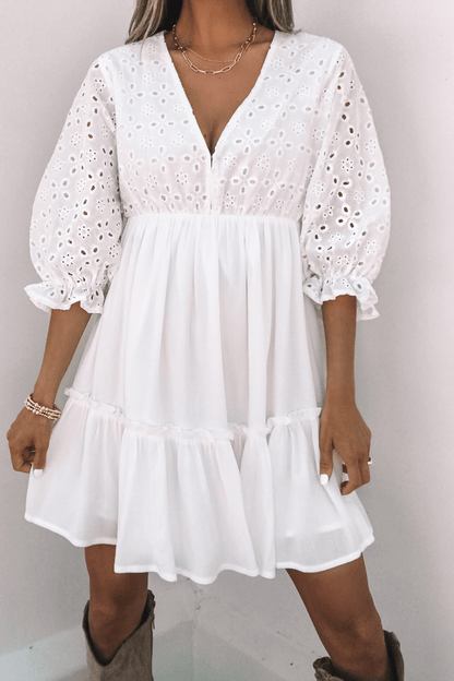 A woman wears the Chloe White Eyelet Tiered Mini Dress with a V-neck, puff sleeves, gold jewelry, and an above-the-knee skirt.