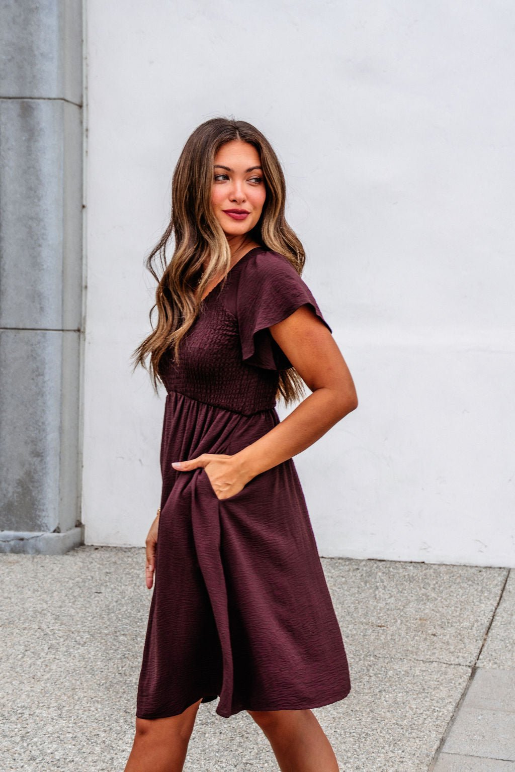 Woman in a Chocolate Brown Smocked Mini Dress - FINAL SALE stands on the sidewalk, one hand in her pocket, looking to the side.