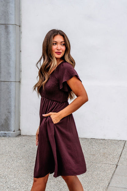 Woman in a Chocolate Brown Smocked Mini Dress - FINAL SALE stands on the sidewalk, one hand in her pocket, looking to the side.