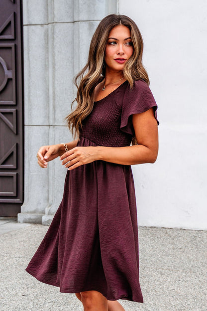 A woman in the Chocolate Brown Smocked Mini Dress - FINAL SALE stands outdoors by a white wall and gray door.