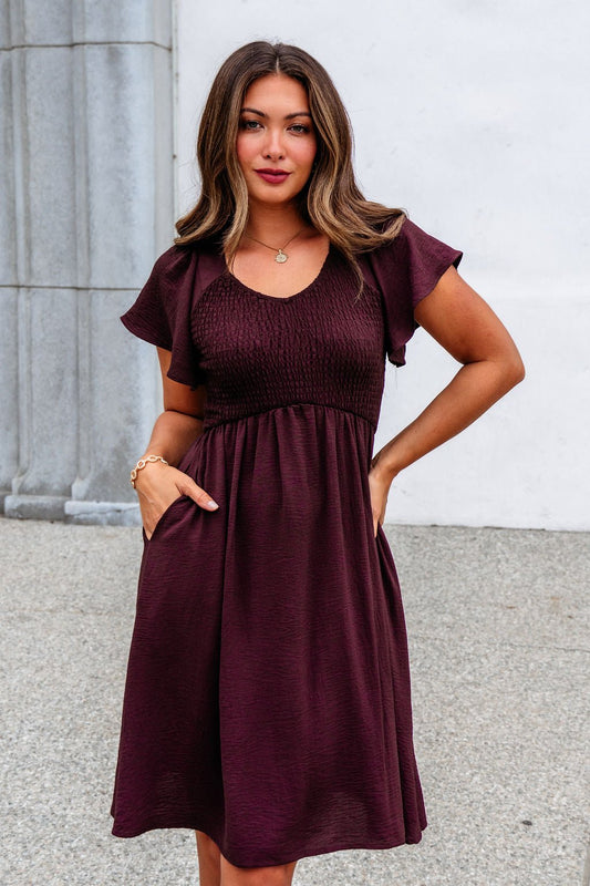 Woman wearing the Chocolate Brown Smocked Mini Dress with a shirred bodice, outdoors, one hand in her pocket.
