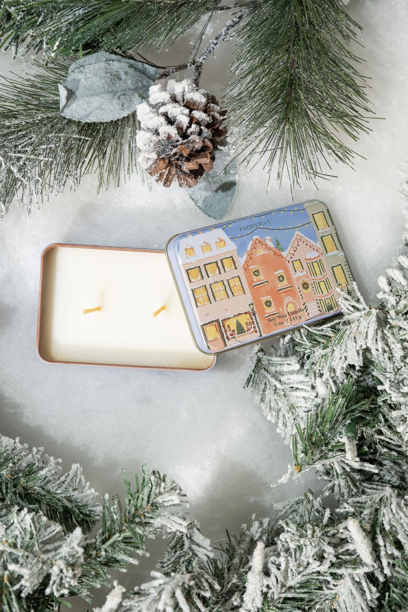 The Christmas Village Print Tin Soy Candle with two wicks sits on snow with frosted pine—perfect festive decor.