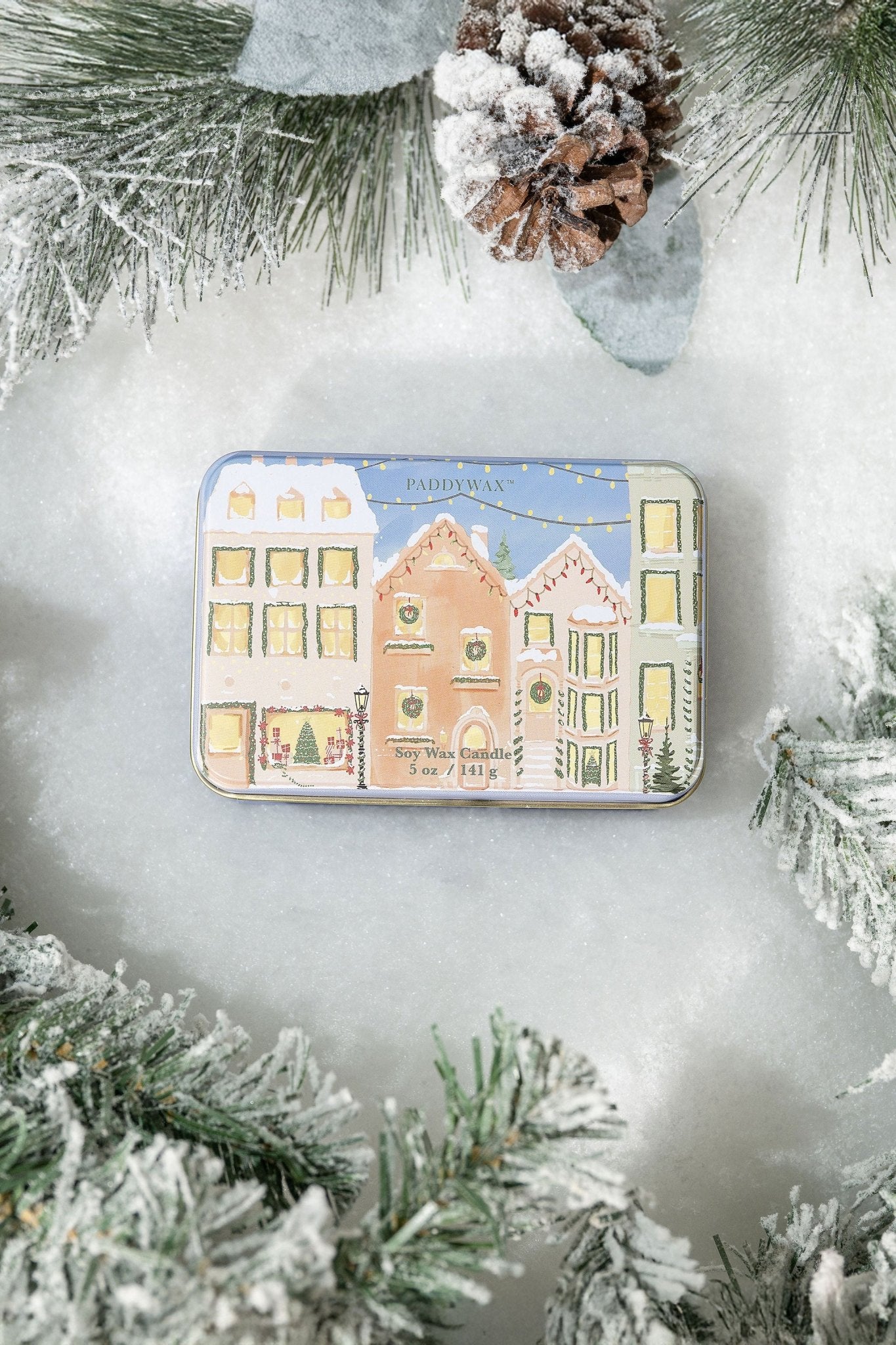 The Christmas Village Print Tin Soy Candle features pastel houses on snow, with frosted pine and a pinecone for a festive touch.