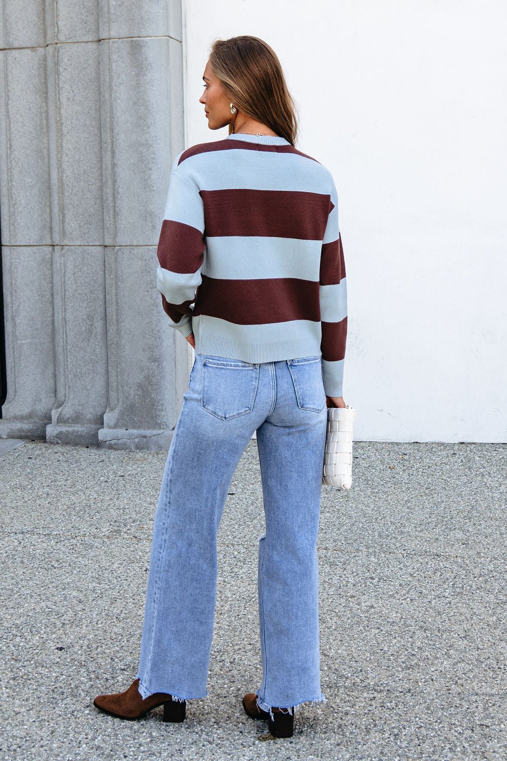 Woman outdoors in a Chunky Brown and Blue Striped Sweater, light blue jeans, boots, white clutch, facing away from the camera.