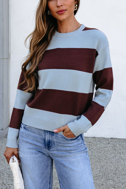 Woman wears chunky brown and blue striped sweater, blue jeans, and holds a white clutch against a light wall.
