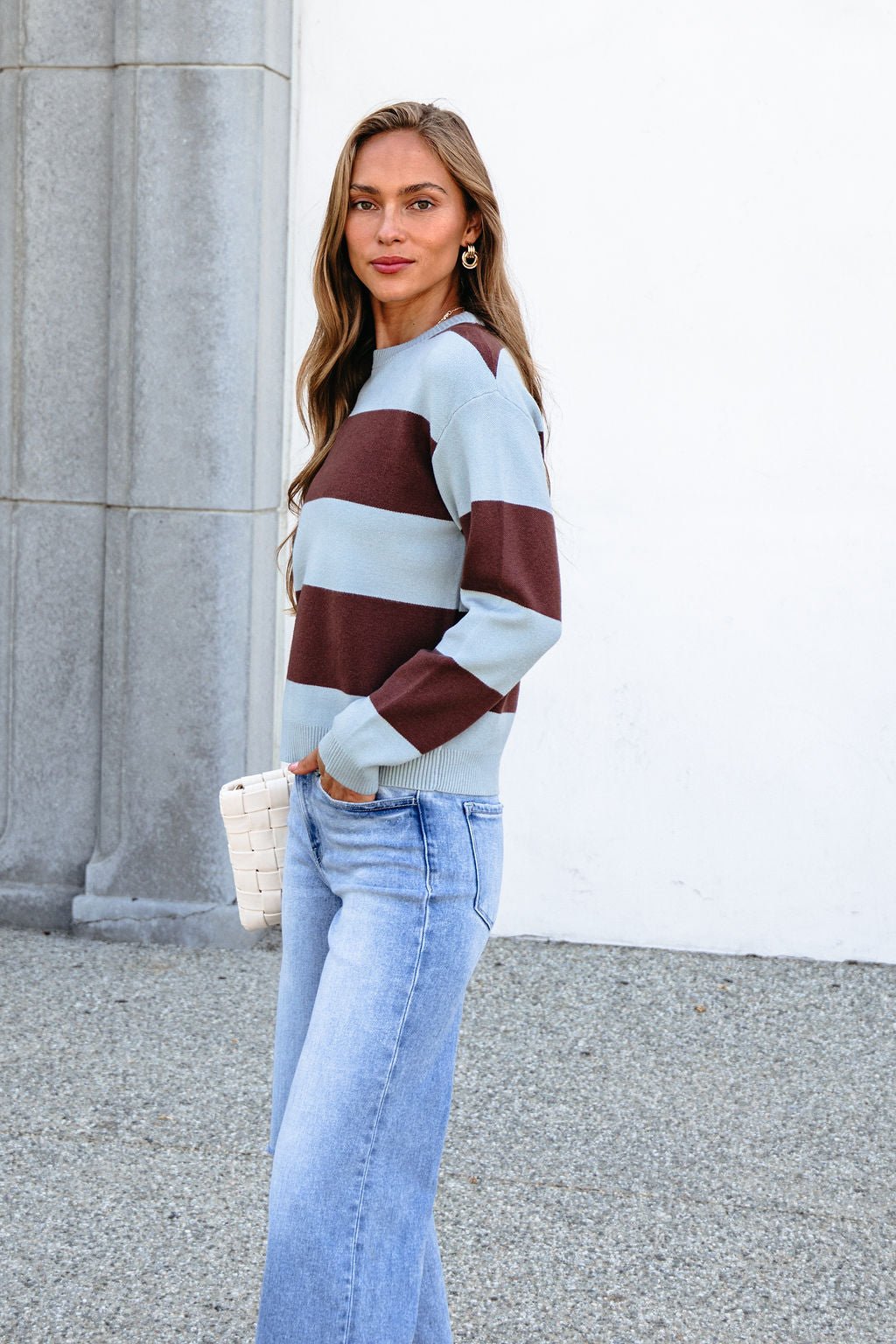 Woman in chunky brown and blue striped sweater and jeans stands outdoors with a white clutch, neutral wall and stone behind her.