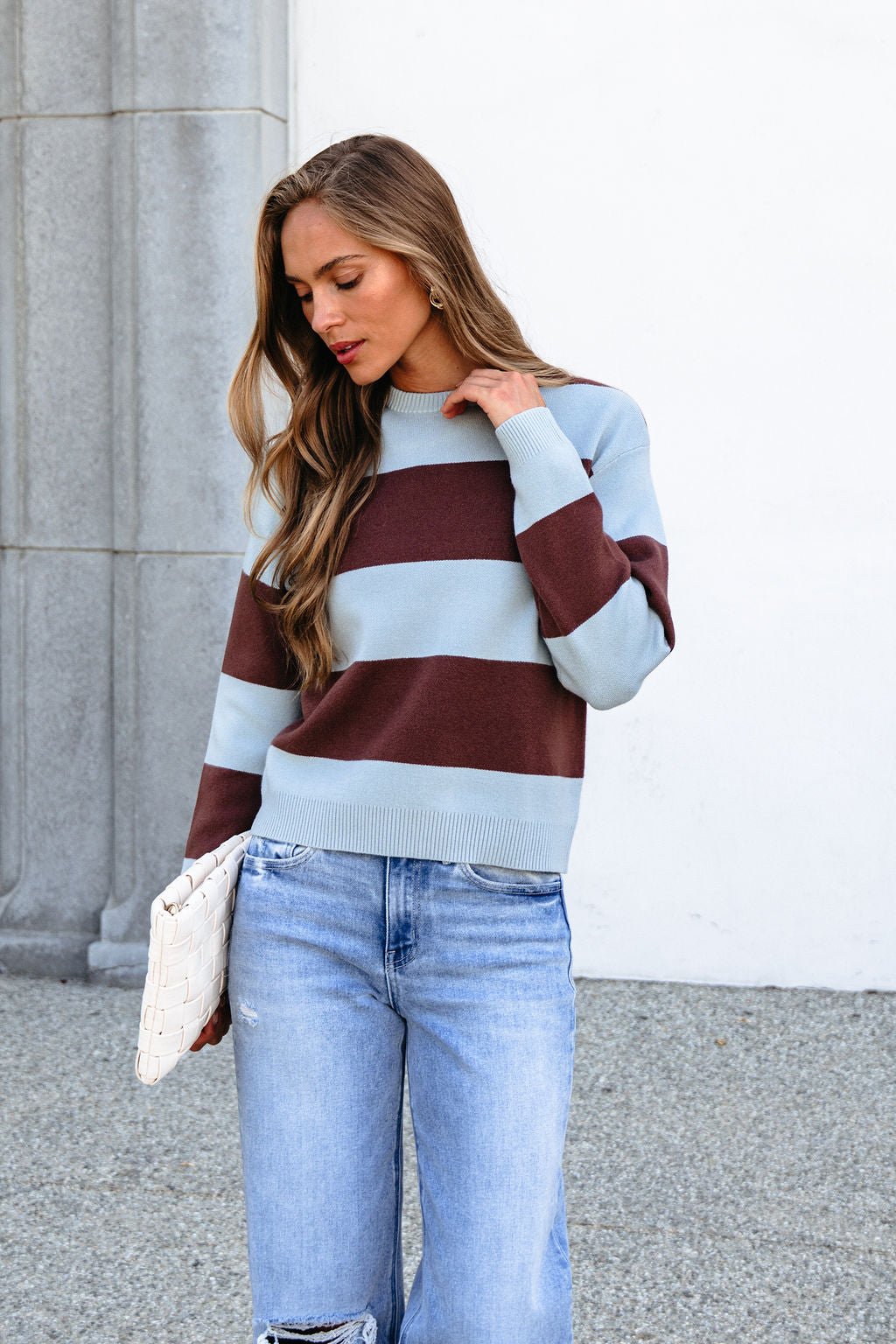 Woman in a chunky brown and blue striped sweater, light blue jeans, holding a white clutch outdoors.