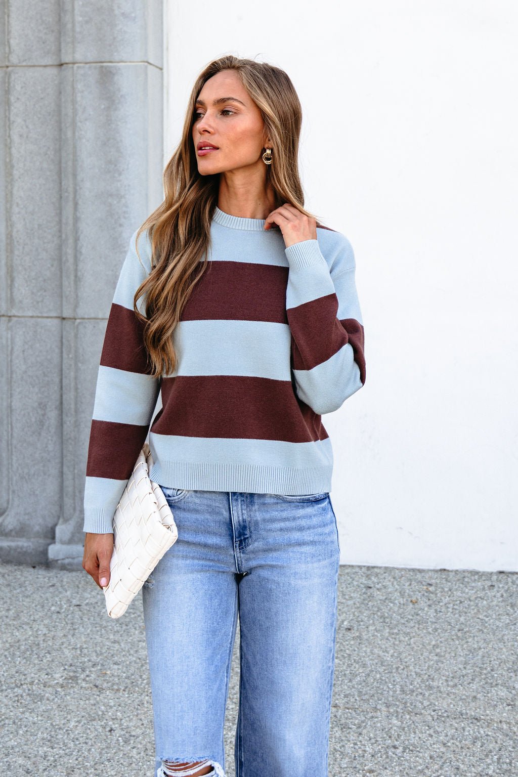 Woman in a Chunky Brown and Blue Striped Sweater, blue jeans, holding a white clutch outdoors by a stone column.