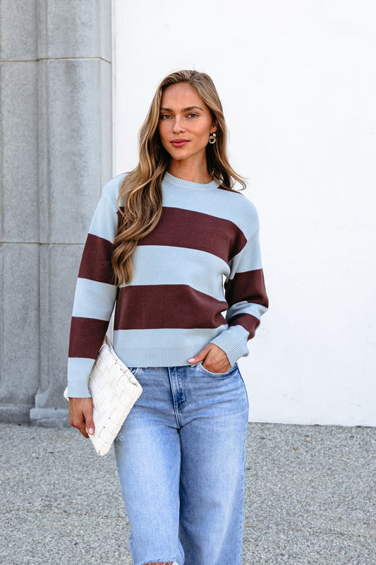 Woman in chunky brown and blue striped sweater and jeans holds a white clutch outside, with long hair against a neutral background.