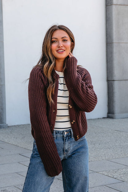 A woman smiles outdoors in a Chunky Brown Button Down Cardigan, striped shirt, and blue jeans against a light wall.