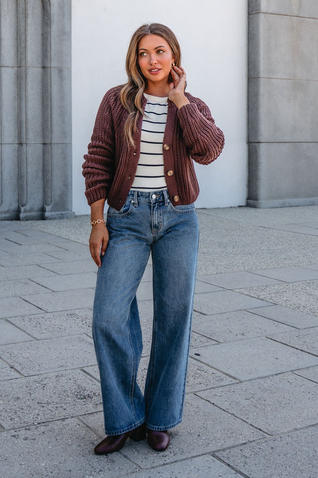 Woman outside in a Chunky Brown Button Down Cardigan, striped top, wide-leg jeans, and brown shoes, touching her hair.