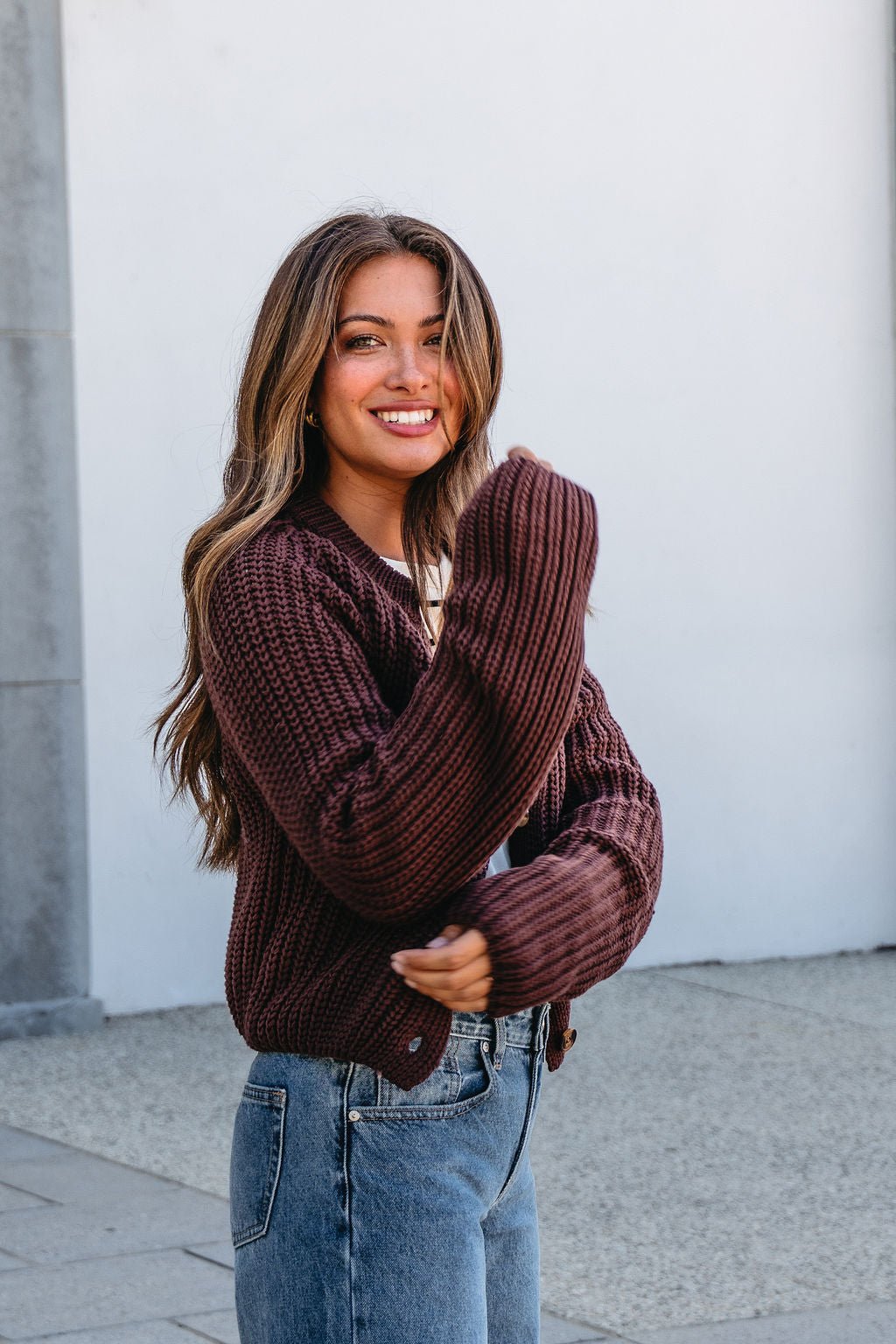 Smiling woman in a Chunky Brown Button Down Cardigan and jeans stands outdoors with her long hair loose.
