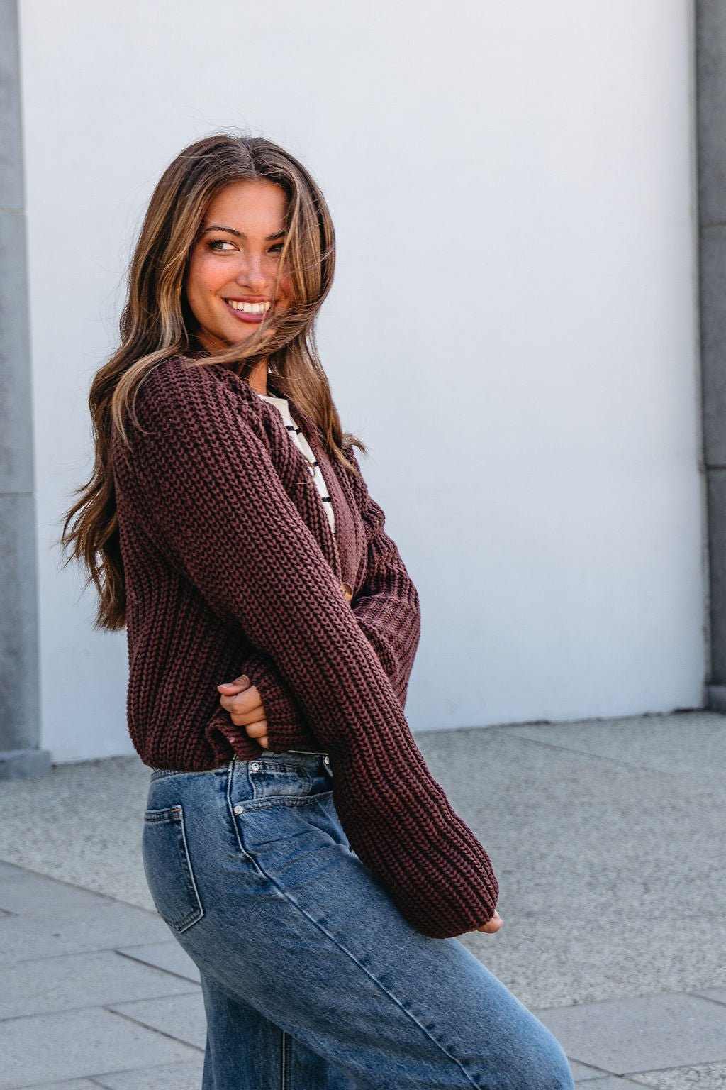 Woman with long brown hair smiles outdoors in a Chunky Brown Button Down Cardigan from Magnolia Exclusive against a light wall.
