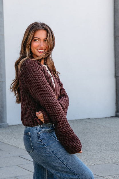 Woman with long brown hair smiles outdoors in a Chunky Brown Button Down Cardigan from Magnolia Exclusive against a light wall.