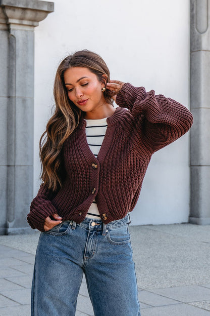 A woman in a Chunky Brown Button Down Cardigan and striped top poses outdoors by a stone and white wall, touching her hair.