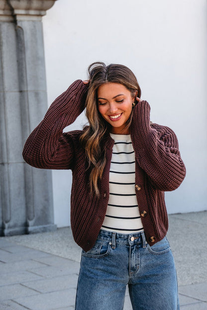 Smiling woman wears a Chunky Brown Button Down Cardigan, striped shirt, and jeans, standing outdoors and touching her hair.
