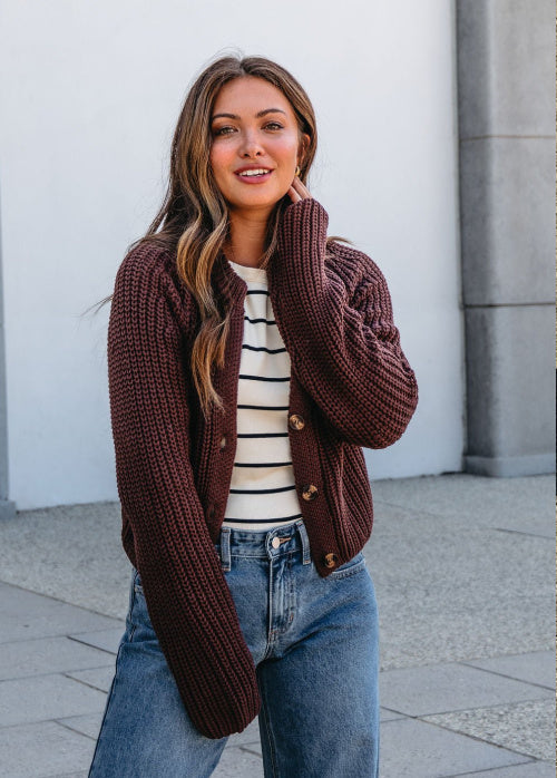 Woman wearing a brown cardigan over a striped shirt with blue jeans, standing against a light-colored wall.