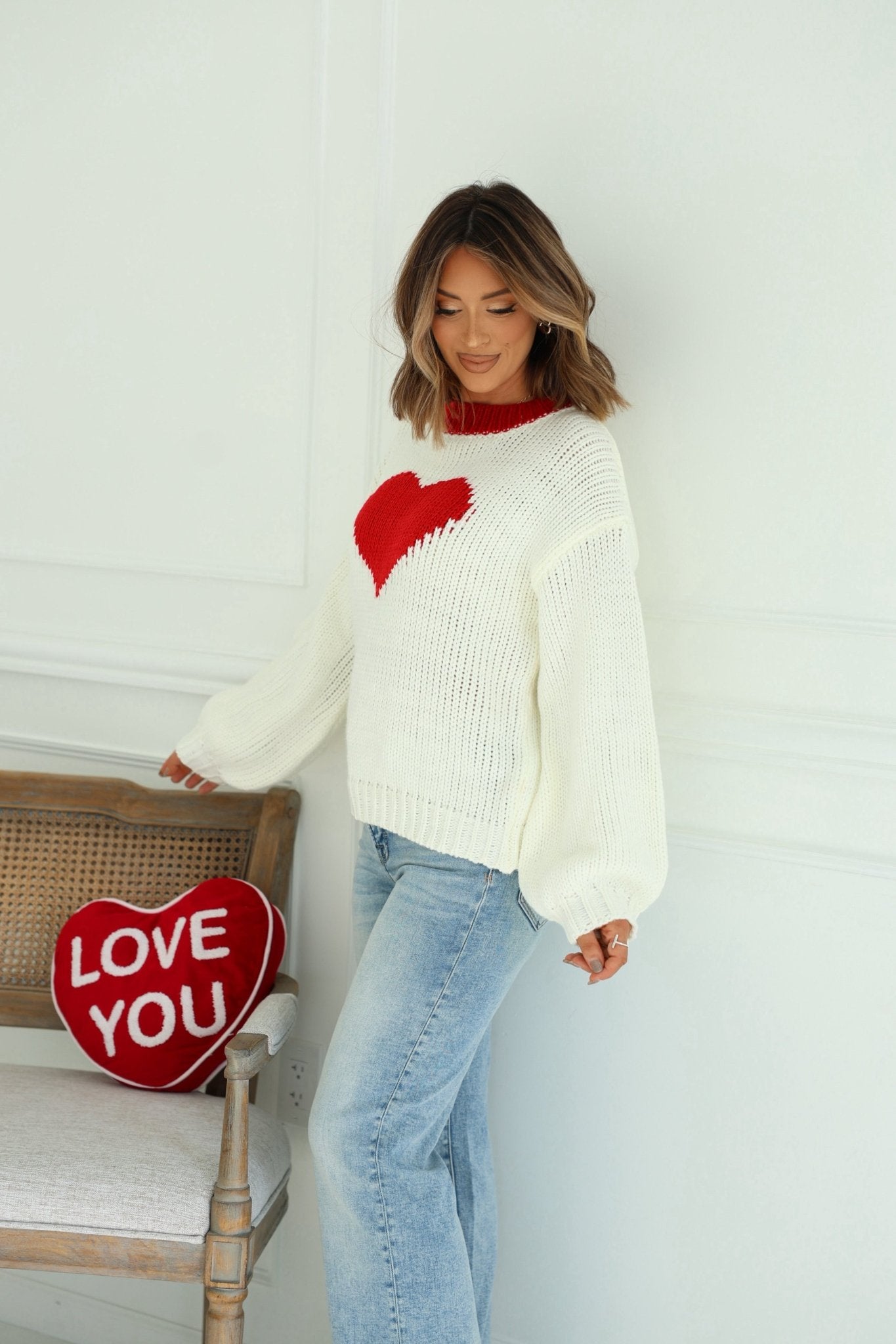 Woman wears the Chunky Cream Heart Shape Sweater by a wooden chair with a LOVE YOU pillow, against a white wall.