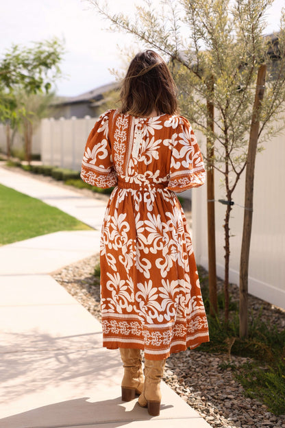 A woman in the Cider Lane Caramel Print Midi Dress walks along a garden path, facing away from the camera.